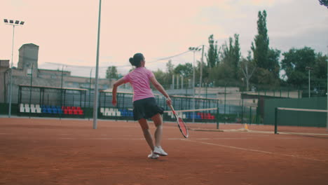 Young-woman-walking-through-tennis-court-with-racket.-Backside-view-of-attractive-brunette-female-in-pink-shirt-and-black-shorts-entering-tennis-hardcourt.-Full-length-follow-shot-with-copy-space